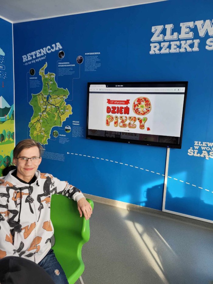 Uczeń pozuje do zdjęcia na tle tablicy multimedialnej.