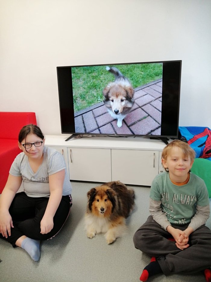 Uczniowie siedzą na podłodze a między nimi waruje pies. 